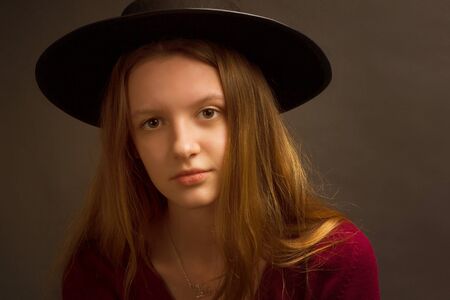 Studio portrait of young blonde girl in a black hat, red blouse on a brown background.の写真素材
