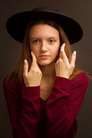 Studio portrait of young blonde girl in a black hat, red blouse on a brown background.の写真素材