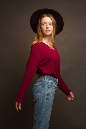 Studio portrait of a young blonde girl in a black hat, red blouse and blue jeans on a brown background.の写真素材