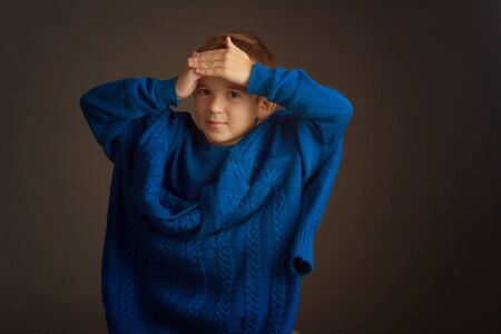 Studio portrait of a dark-haired boy with a short haircut in a blue t-shirt and a knitted sweater, draped over his shoulders, holding his hands on his forehead, smiling. On a brown backgroundの写真素材