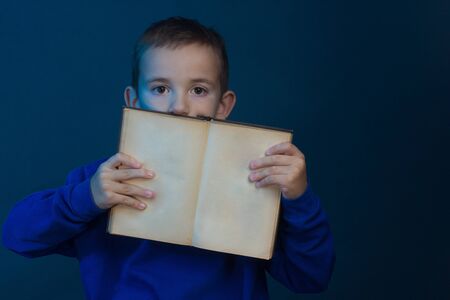 Studio portrait of a boy on a blue background in a classic t-shirt. he holds an open book in his hands, with empty pages - a copyspace to the viewer.の写真素材