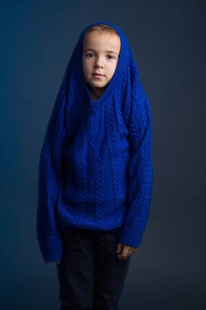 Studio portrait of dark-haired boy with a short haircut in a blue classic knitted sweater pulled playfully over his head. The concept of play, pampering and joy.の写真素材