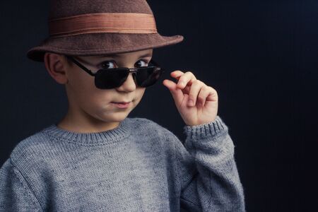 Studio portrait of a boy in a grey sweater, brown hat and sunglasses, on black background, holding his glasses with his handの写真素材