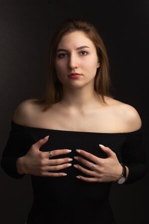 Portrait in the photo Studio of a brown-haired girl, on a black background in black clothes with open shoulders. Short hair.. Hands held on the chestの写真素材