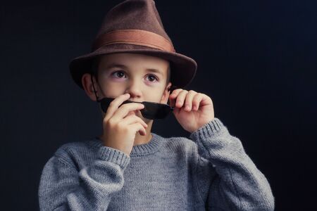 Studio portrait of a boy in a grey sweater, brown hat and removes glasses at nose level with his hands, on black backgroundの写真素材