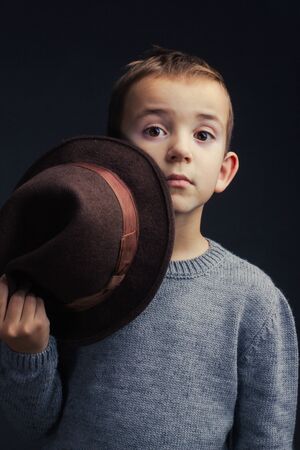 Studio portrait of a boy in a gray sweater, on a black background, holding a brown hat behind the brim, hiding and peeking out from behind itの写真素材