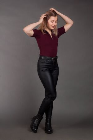growth Portrait of the blonde in the red sweater with short sleeves, leather trousers and boots. Studio shot on dark gray background. hands at the head of the hair, bent right kneeの写真素材