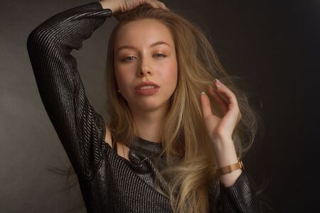 portrait of a blonde in the Studio on a dark uniform background in a black shiny blouse with open shoulders. closed eyes, open mouth and hands straightens hairの写真素材