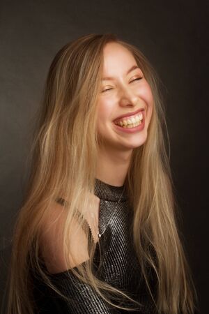 portrait of a blonde in the Studio on a dark uniform background in a black shiny blouse with open shoulders. laughing with his eyes closedの写真素材