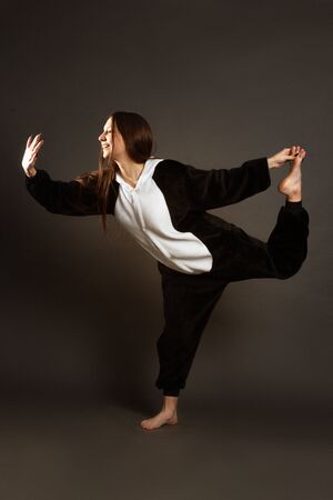 a portrait of growth of a brunette girl in the Studio on a plain black background in black and white pajamas with a hood, jumps, standing on one leg, the other behind, holding her handの写真素材