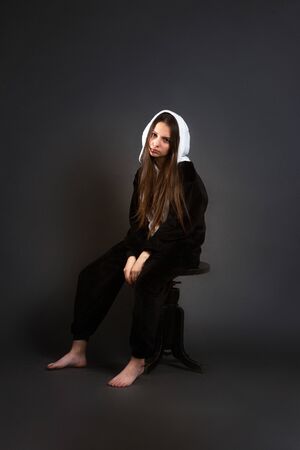 a portrait of growth of a brunette girl in the Studio on a plain black background in black and white pajamas with a hood, sitting on a chair, sadの写真素材