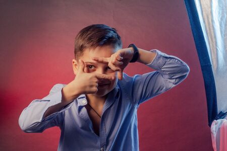 The Studio portrait of boy is a brunette, in a blue shirt on a purple and maroon background. Framing Face With Fingersの写真素材