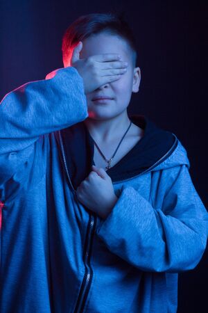 Studio portrait of a dark-haired boy, on a dark background, in a hoodie jacket in the hood. light color: red and blue. covers his face with his handの写真素材