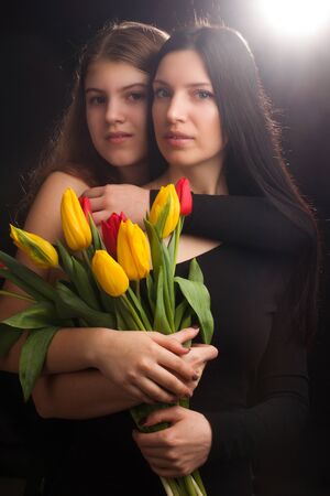 Portrait of girl and woman, mother and daughter, in a black one-shoulder dress open, naked , with red and yellow tulips, in Studio on black background, backlit and lens flareの写真素材