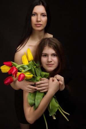 Portrait of girl and woman, mother and daughter, in a black one-shoulder dress open, naked , with red and yellow tulips, in Studio on black backgroundの写真素材