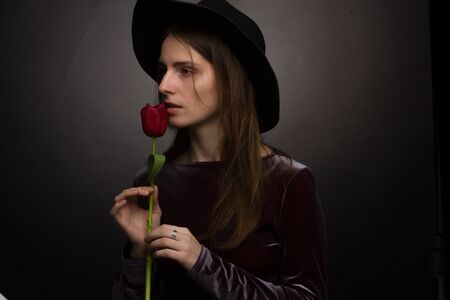 The girl in the purple dress and wide-brimmed black hat on a dark background in the Studio with a red Tulip flower in the hand . creative portrait of a sensual womanの写真素材