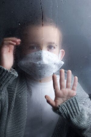 Boy medical face mask, looking through the glass, laying a hand on him. The concept of isolation and self-isolation during an outbreak of coronavirusの写真素材