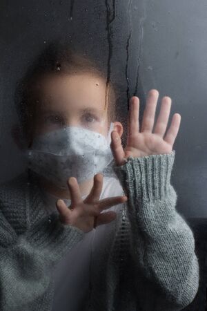 Boy medical face mask, looking through the glass, laying a hand on him. The concept of isolation and self-isolation during an outbreakの写真素材