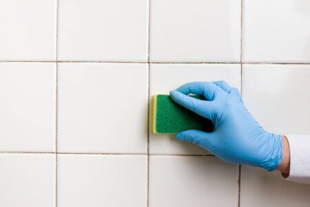 a hand in a blue rubber glove holds a yellow-green microfiber cleaning sponge, washing a white tile...