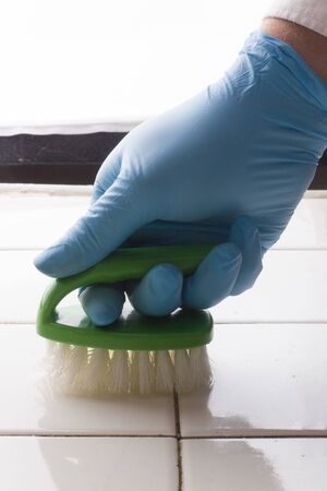 a hand in a blue rubber glove holds a green cleaning brush, washing the white tile surface