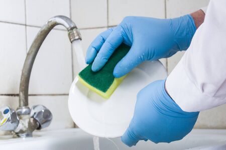 blue gloved hands wash a white plate over the kitchen sink against a white tiled wallの写真素材