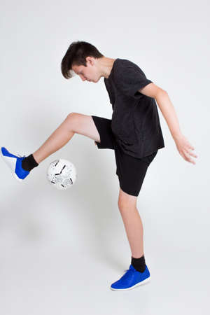 A young man in a black sports t-shirt and shorts, blue sneakers, trains and plays with a soccer ball. Isolated on white.の写真素材