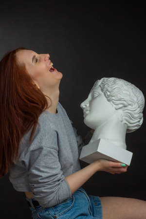 Red-haired young girl on black background in a gray T-shirt, holding a plaster bust