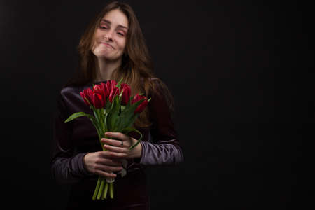 Studio portrait of a young woman on a black background with a bouquet of red tulips in her handsの写真素材