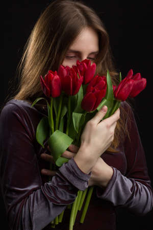 Studio portrait of a young woman on a black background with a bouquet of red tulips in her handsの写真素材