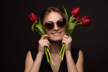 Studio portrait of a young woman in sunglasses, on a black background, with a bouquet of red tulips...