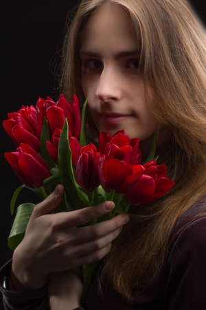 Studio portrait of a young woman on a black background with a bouquet of red tulips in her handsの写真素材