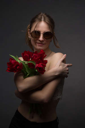 Studio portrait of a young woman in sunglasses, on a black background, with a bouquet of red tulips in her handsの写真素材