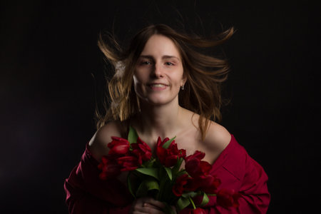 Studio portrait of a young woman on a black background with a bouquet of red tulips in her handsの写真素材