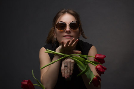Studio portrait of a young woman in sunglasses, on a black background, with a bouquet of red tulips...