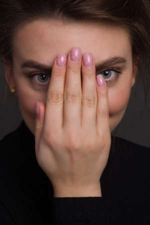 Young brunette woman, with gathered hair hairstyles, in the studio on a black background in a black tight-fitting bodysuit. he covers his face with his handの写真素材