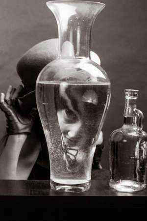 black and white A brunette woman with long hair, wearing a beige blouse, brown hat and gloves on a gray studio background. Looks at the camera through a glass vaseの写真素材