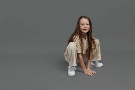 Studio portrait of a young girl with brown hair, with long hair, on a gray background in beige clothes. Squatting on the groundの写真素材