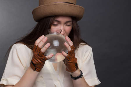 A brunette woman with long hair, wearing a beige blouse, brown hat and gloves on a gray studio background. Looks at the camera through a glass vaseの写真素材