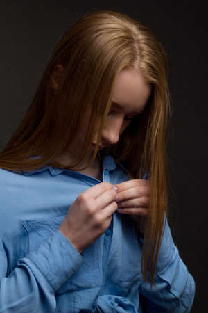 A young woman in a blue blouse, a portrait on a black studio background. Fastens a button on her shirt.の写真素材