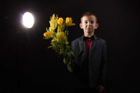 a boy with vitiligo in a red T-shirt, gray jacket and bow tie, with a bouquet of mimosa and yellow tulipsの写真素材