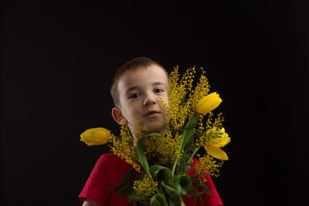 a boy with vitiligo in a red T-shirt with a bouquet of mimosa on a black studio backgroundの写真素材