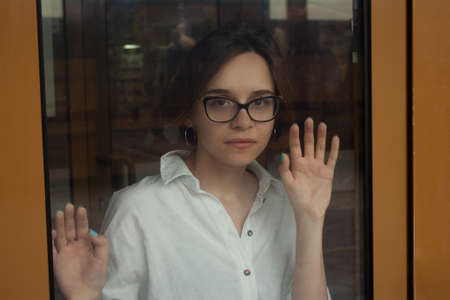 A young woman with short brown hair, wearing a white shirt, with glasses, behind the glass