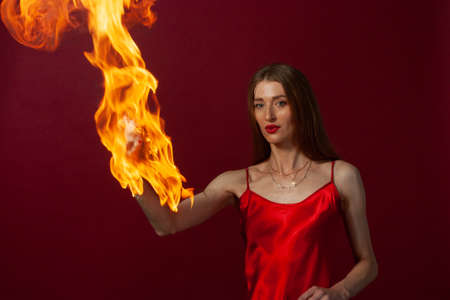 A young woman with long brown hair, in a red dress on a red background playing with fireの写真素材