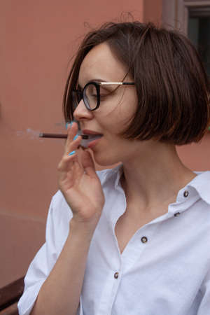 A young woman with short brown hair, wearing a white shirt, with glasses, smokes a cigaretteの写真素材
