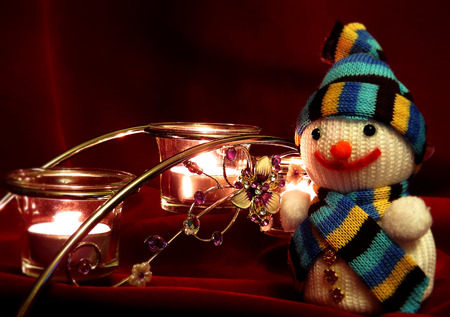 Happy new year or Merry Christmas card with snowman. Knitting from colored thread. Beautiful candles burning in the evening. On dark red fabric background.の写真素材