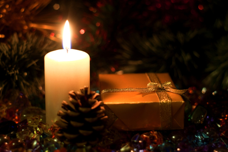 Christmas and New Year`s evening burning candle and present gift box wrapped in paper with bows of gold ribbons. Retro style greeting card yellow toned background with holiday tinsel and fir cone.の写真素材