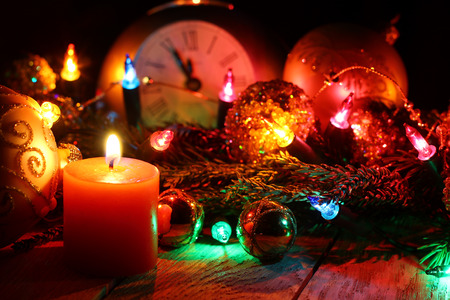 New Year`s card with Christmas tree fir branches, midnight clock, burning candle, golden pine cones, balls, garland lights on vertical old wooden desk table background. Big copyspace for holiday congratulations.の写真素材