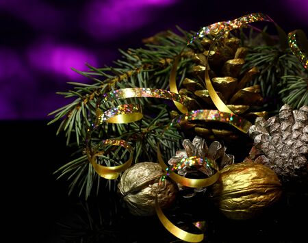 Christmas gold decorations with fir tree brunch, golden ribbons, walnuts and pine cones  on a black mirror reflection surface and dark purple bokeh background with snowflakes. New year`s  card.の写真素材