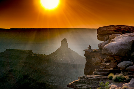 Lone man sitting on a ledge overlooking the desert canyon landscape during a golden sunsetの写真素材