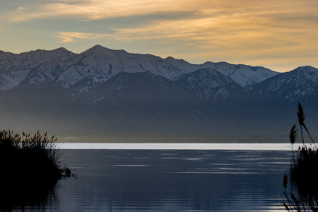 Lake with grass along the shoreline against a backdrop of snow-capped mountains during a golden sunsetの写真素材